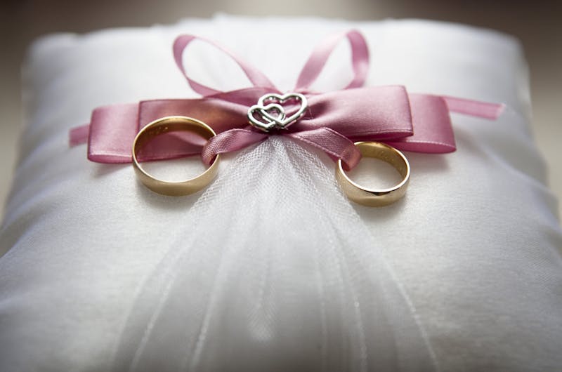 Close-up of elegant wedding rings on a silk pillow with a pink ribbon and a heart decoration, symbolizing love.